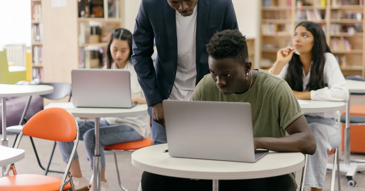 Students working on laptops in a library while a tutor helps one of them