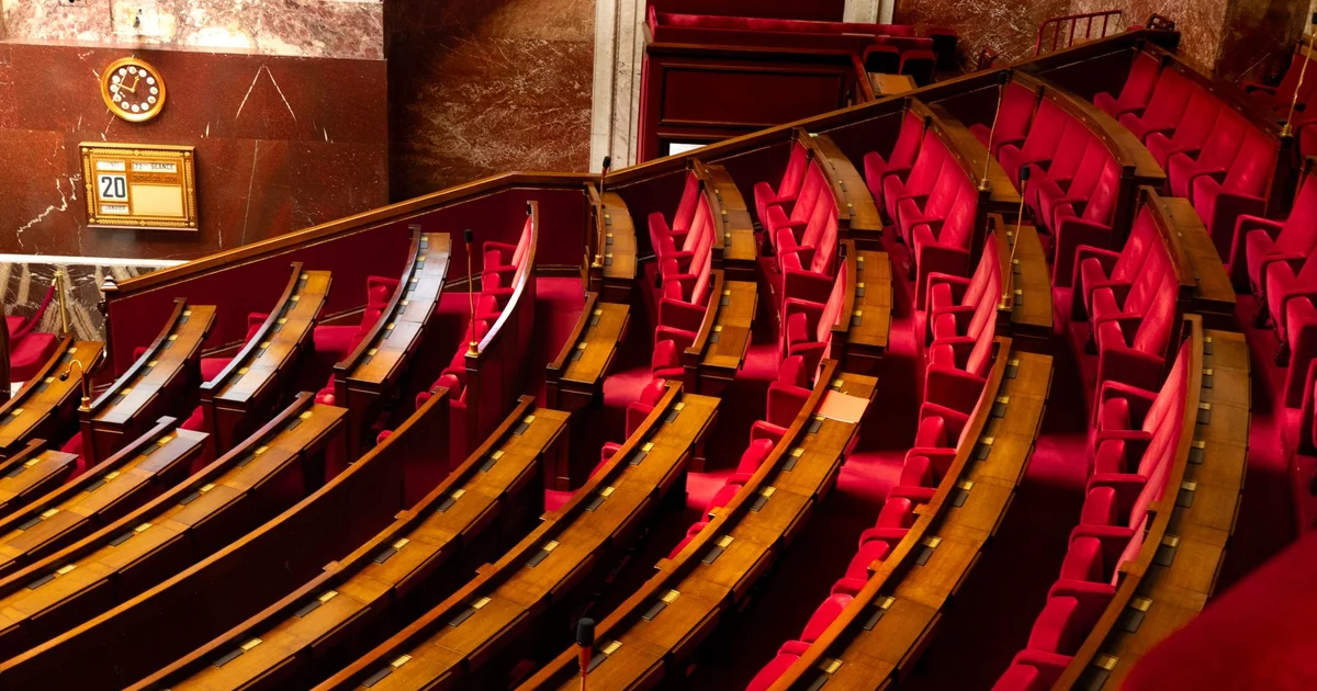 Interior de una cámara legislativa vacía con escritorios de madera y butacas rojas