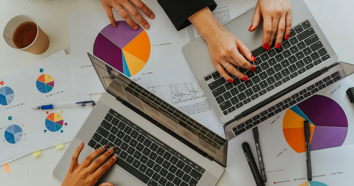 Overhead view of a work table with two laptops, hands typing on the keyboards and printed reports with colorful pie charts, pens and a cup of coffee
