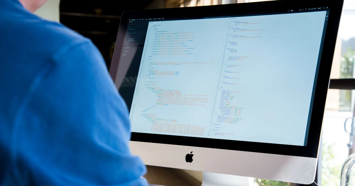 Programador de camisa azul frente a un iMac con un editor de código abierto en la pantalla