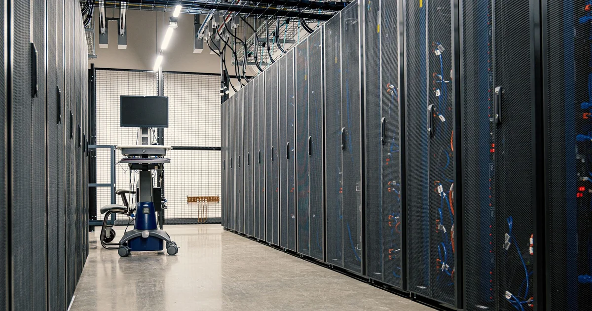 Data center aisle with rows of black server racks with mesh doors and blue and orange cables, and a cart with a monitor in the background