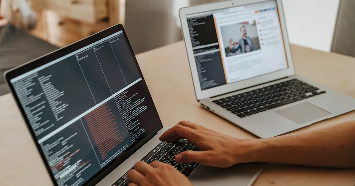 Manos de un programador escriben código en una laptop mientras otra laptop al lado muestra una videollamada