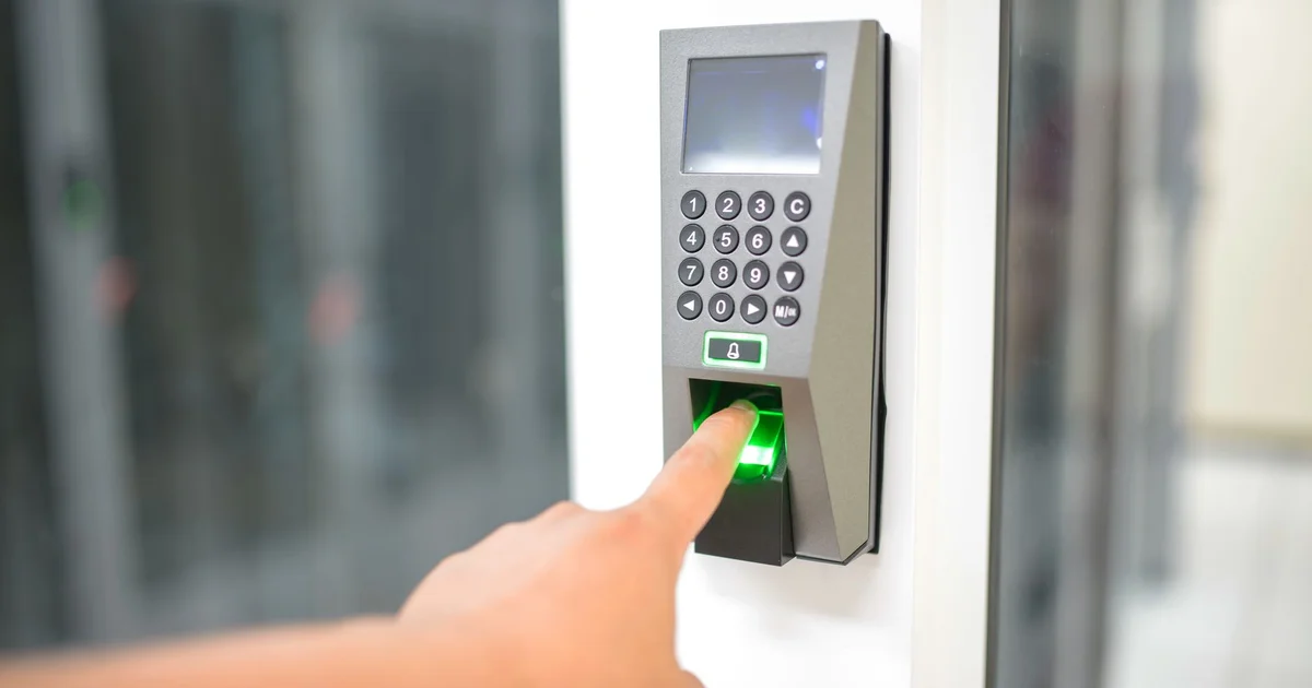 Finger on the green-lit fingerprint reader of an access control terminal with a keypad and screen, next to a glass door