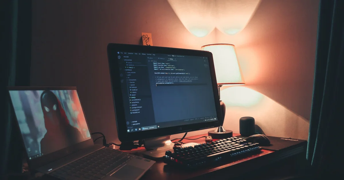 Dim desk with a monitor showing JavaScript code in Visual Studio Code, a backlit keyboard, a lit lamp and, on the left, a laptop displaying a hooded figure