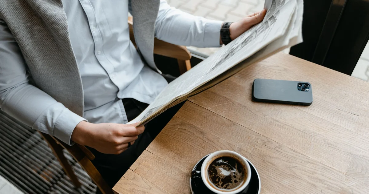 Man reading a newspaper at a café table next to a cup of coffee and his phone