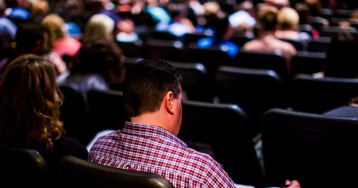 Público sentado en un auditorio durante una conferencia, con un asistente tomando notas en primer plano