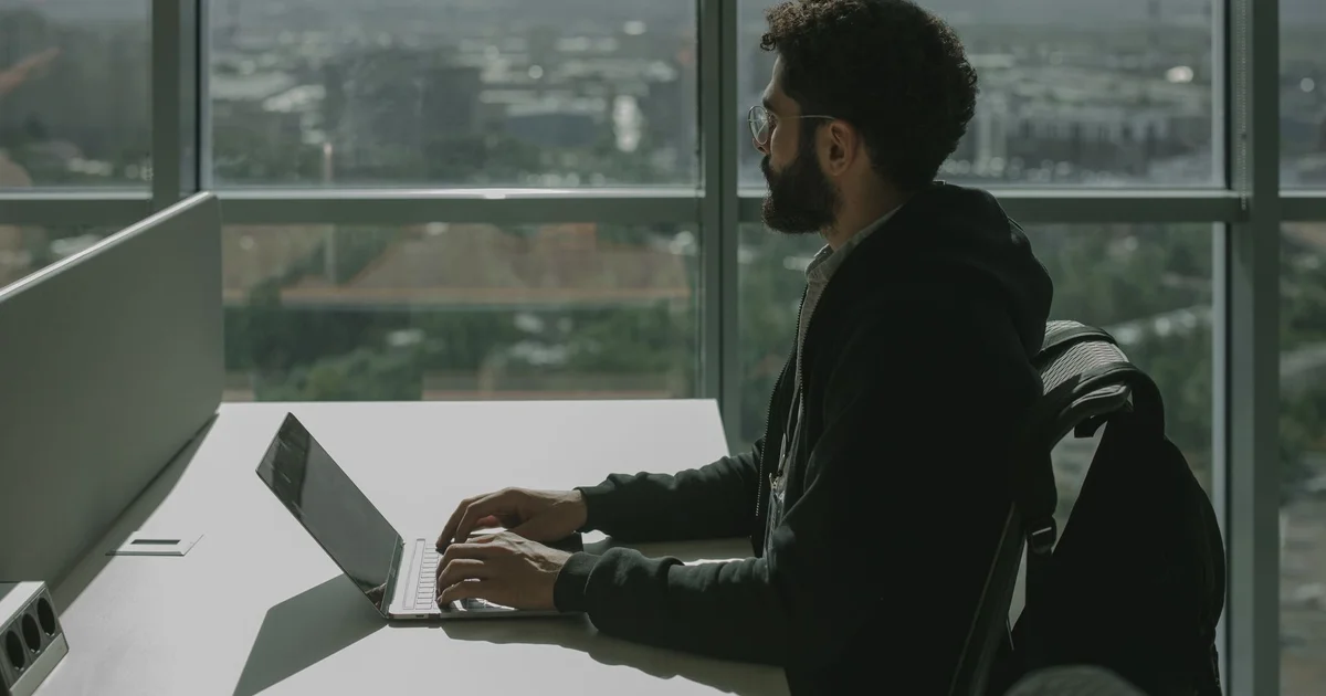 Bearded man with glasses in a black hoodie typing on a laptop at a white office desk in front of a large window overlooking the city